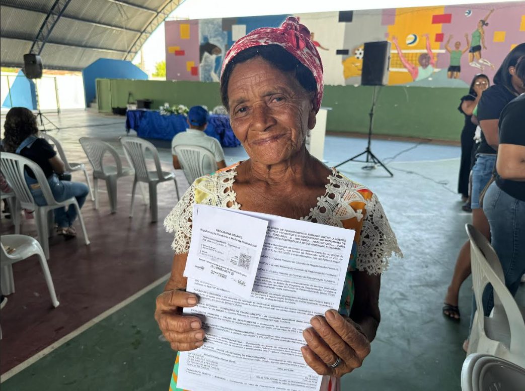 Ao centro senhora mostrando o documento da regularização, em torno pessoas sentadas em quadra